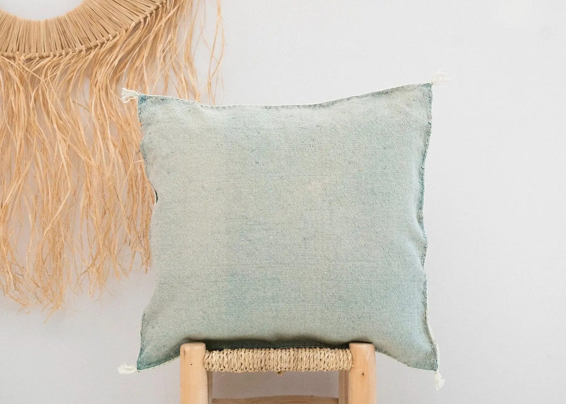 Een lichtblauw vierkant kussen staat op een geweven kruk tegen een witte muur. Een strodecoratie is gedeeltelijk zichtbaar aan de linkerkant.