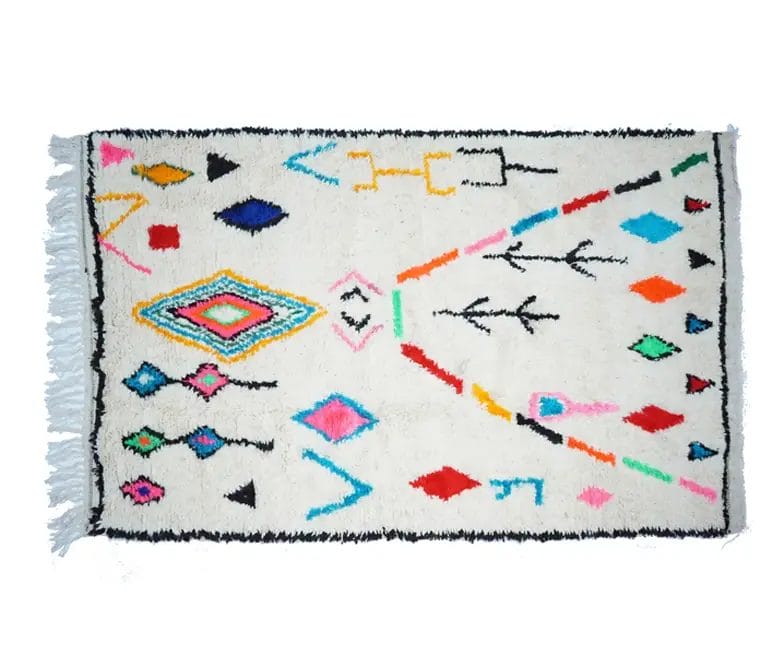 Het Handgemaakt Berber vloerkleed is een rechthoekig vloerkleed met geometrische patronen in verschillende kleuren, zoals blauw, roze, zwart en oranje, op een witte achtergrond, met franjes aan één rand.
