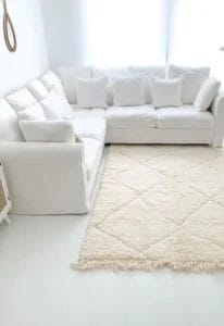 Een witte L-vormige bank met meerdere witte kussens staat in de hoek van een lichte kamer. Een groot gebroken wit, gefranjerd Marokkaans handgemaakt Beniourain Berber wit vloerkleed bedekt de vloer voor de bank.