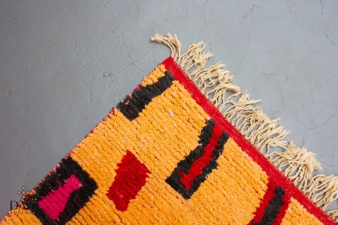 Een hoek van een kleurrijk geweven tapijt met geometrische patronen en franjes ligt op een grijze vloer met textuur.
