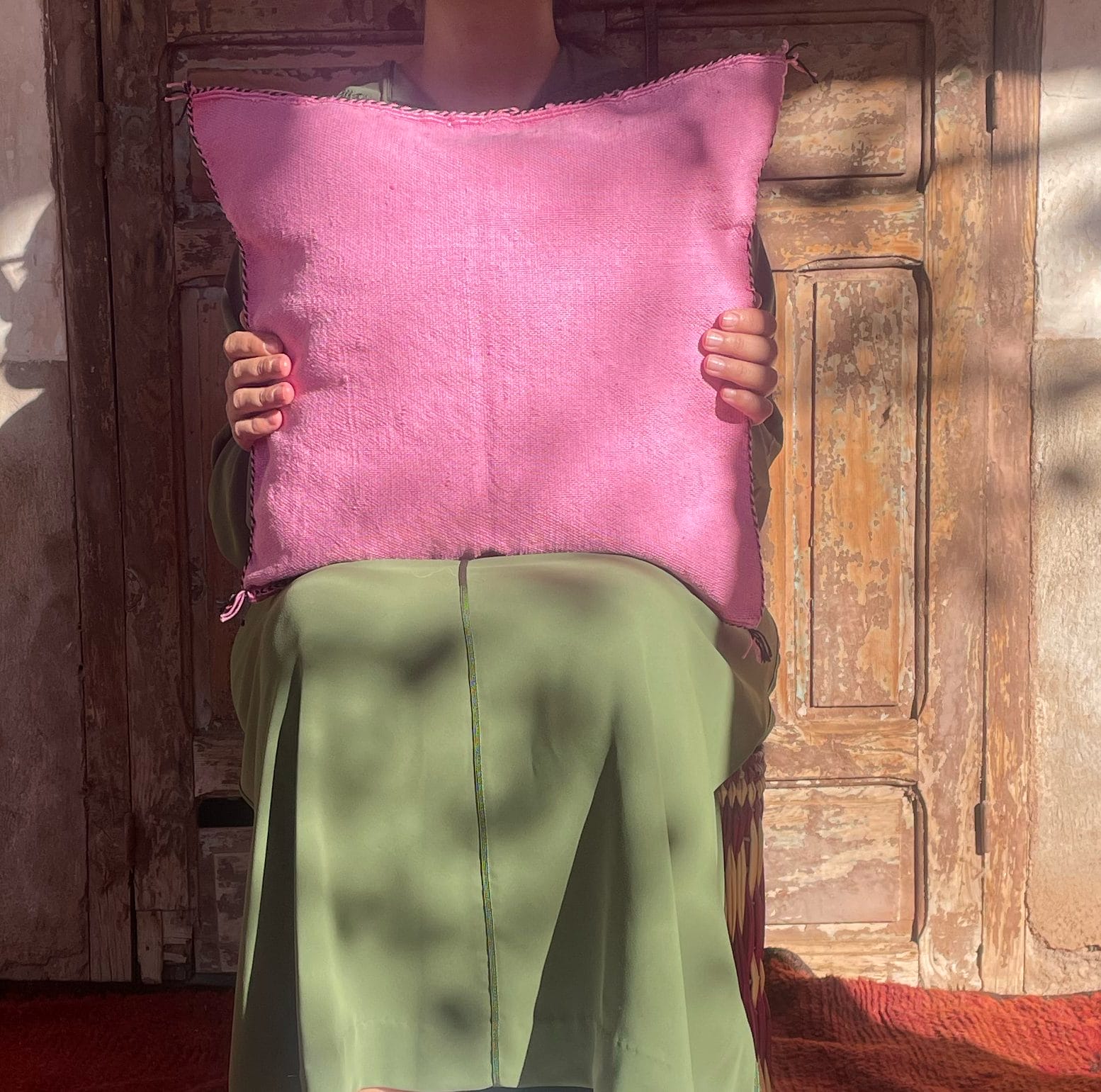 Persoon zittend, met een roze kussen voor zich. Achtergrond toont een verweerd, houten oppervlak.