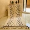 Een handgemaakt Marokkaans Beni Ouarain-kleed met zwarte geometrische ruitpatronen en franjeranden ligt in een betegelde kamer bij een glazen deur. Drie groene decoratieve vazen en twee zwarte ornamenten staan op een houten plank.