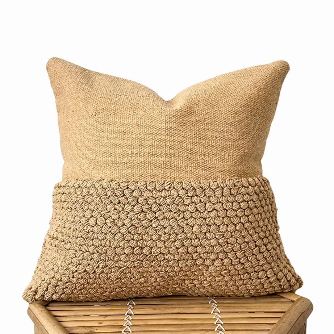 Het Kussenhoes 50 x 50 cm | Amity, een bruin sierkussen met een bovenste helft van gladde stof en een onderste helft met een gestructureerd lusmotief, staat elegant op een bamboe krukje en vormt een mooie aanvulling op het aangrenzende tapijt.