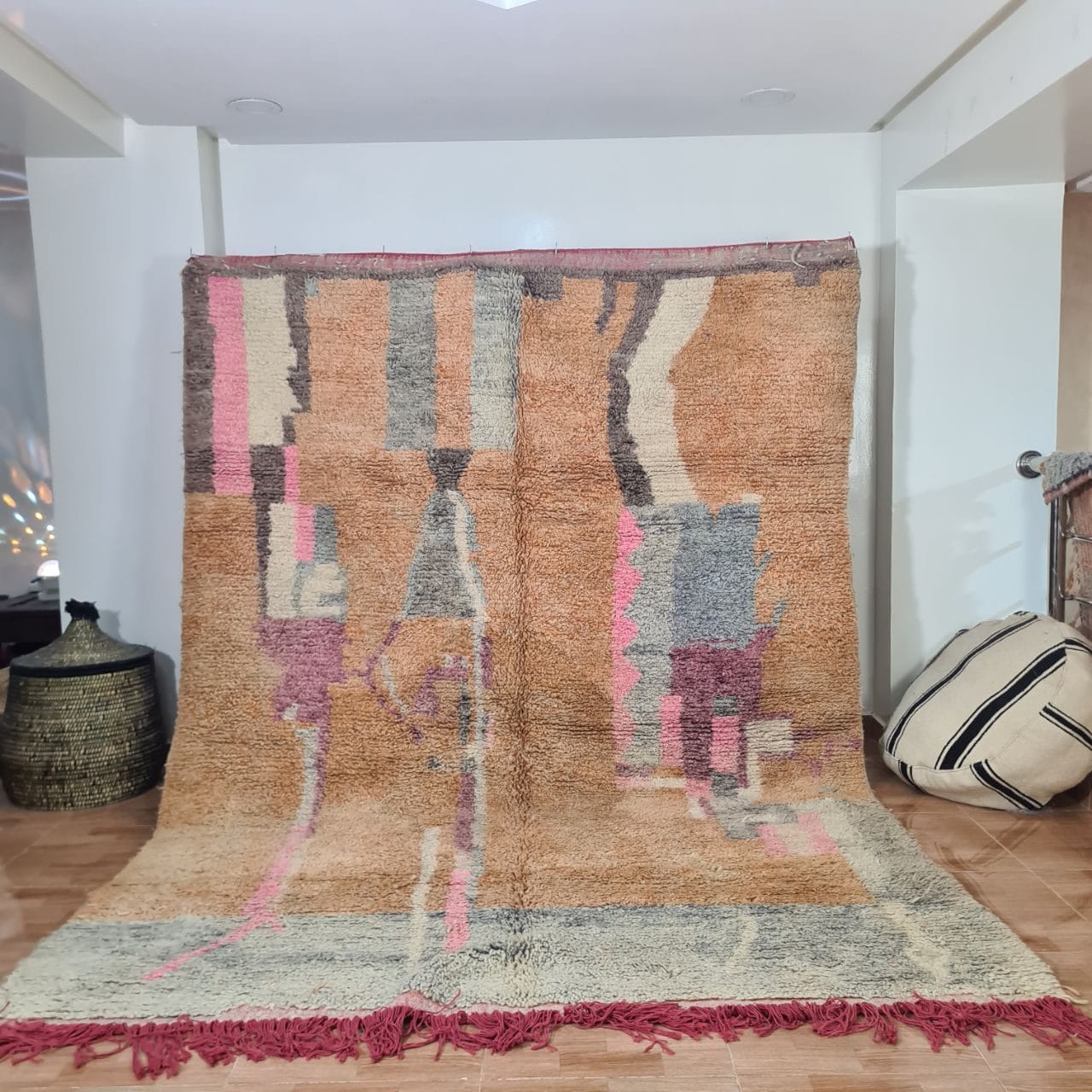 Een grote, abstracte, veelkleurige HANDGEMAAKT MAROKKAANS TAPIJTTAPIJT Boujaad 200 x 300 cm 27 met een mix van beige, roze, grijs en bruintinten staat rechtop in een kamer met beige tegelvloer. Een gevlochten mand en twee gestreepte kussens staan in de buurt.