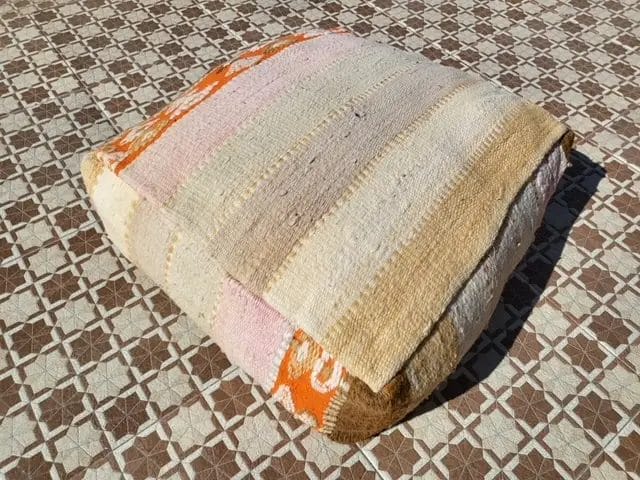 Een vierkant kussen met beige, roze en oranje strepen op een tegelvloer met patroon.