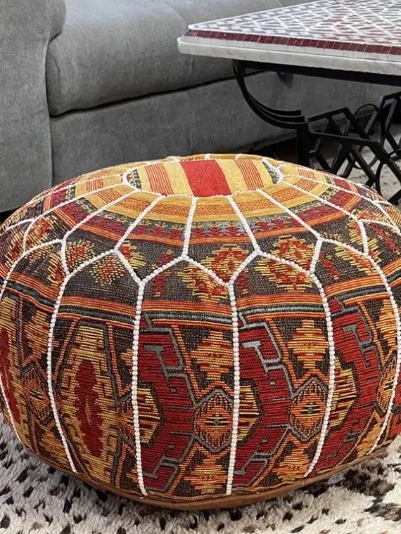Een kleurrijke poef met patroon staat op een gestructureerd tapijt voor een bank en een decoratieve tafel.