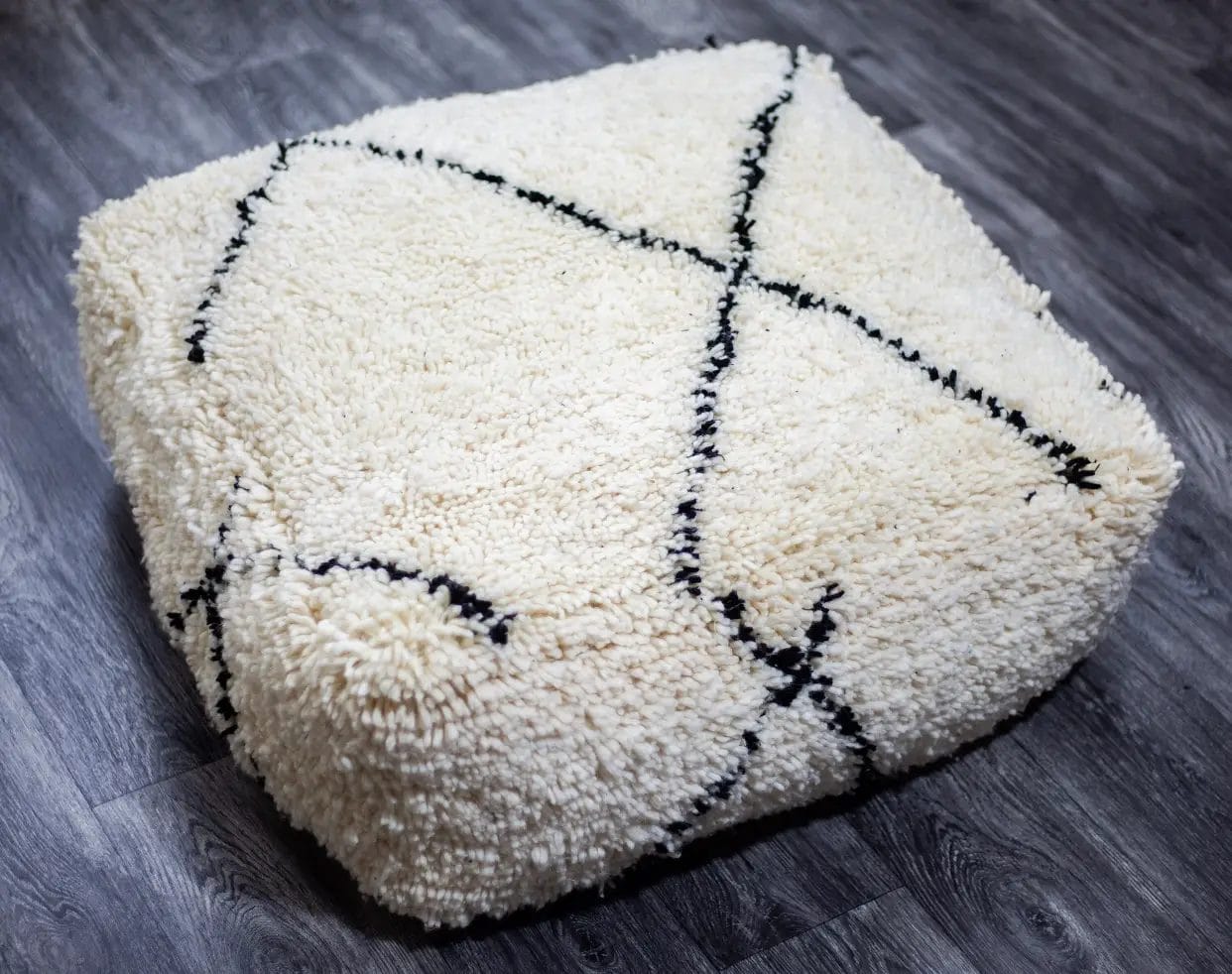 Een vierkante, zachte, witte poef met zwarte kruispatronen staat op een donkere houten vloer.