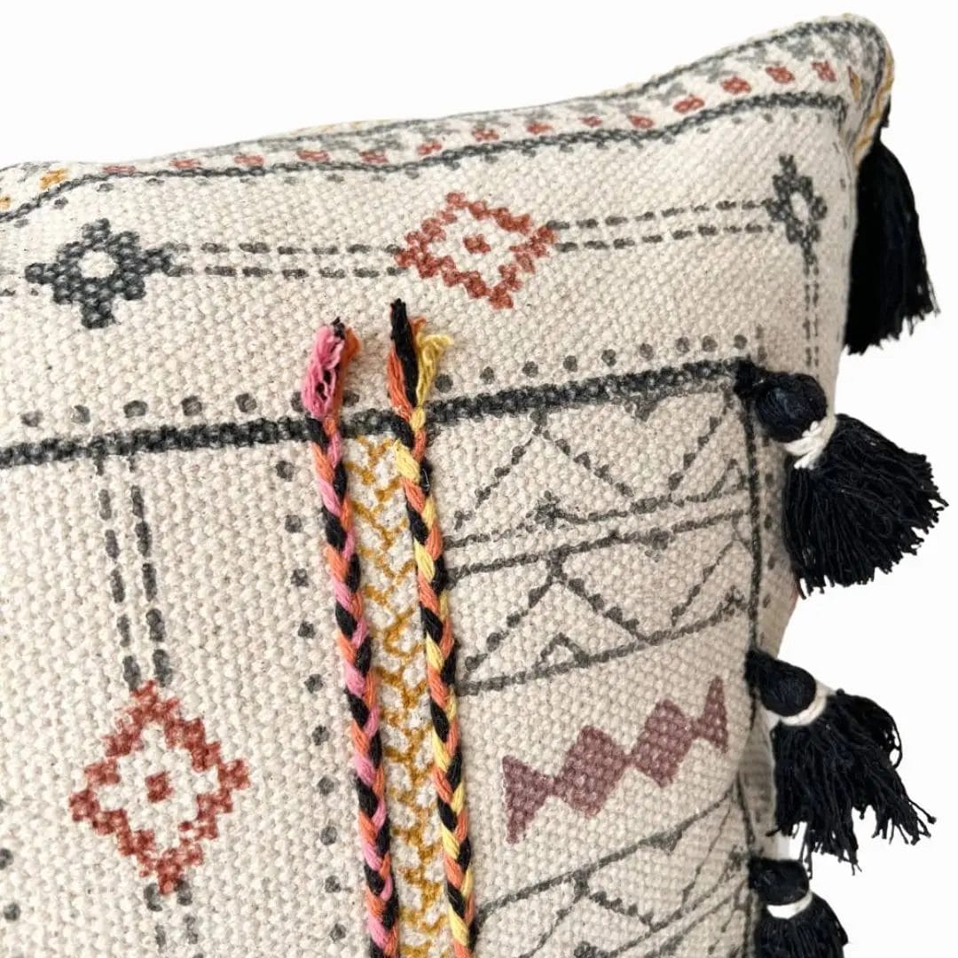 Close-up van een decoratief geweven kussen met geometrische patronen, gevlochten kwastjes in roze, geel en zwart en zwarte kwastjes langs de rand.