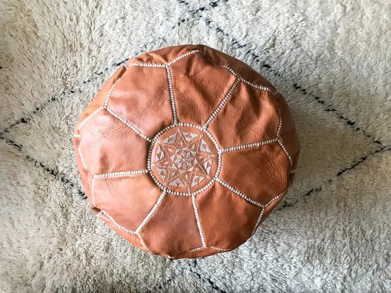 Een ronde, bruine leren poef met decoratieve stiksels aan de bovenkant, geplaatst op een lichtgekleurd vloerkleed met een geometrisch patroon.