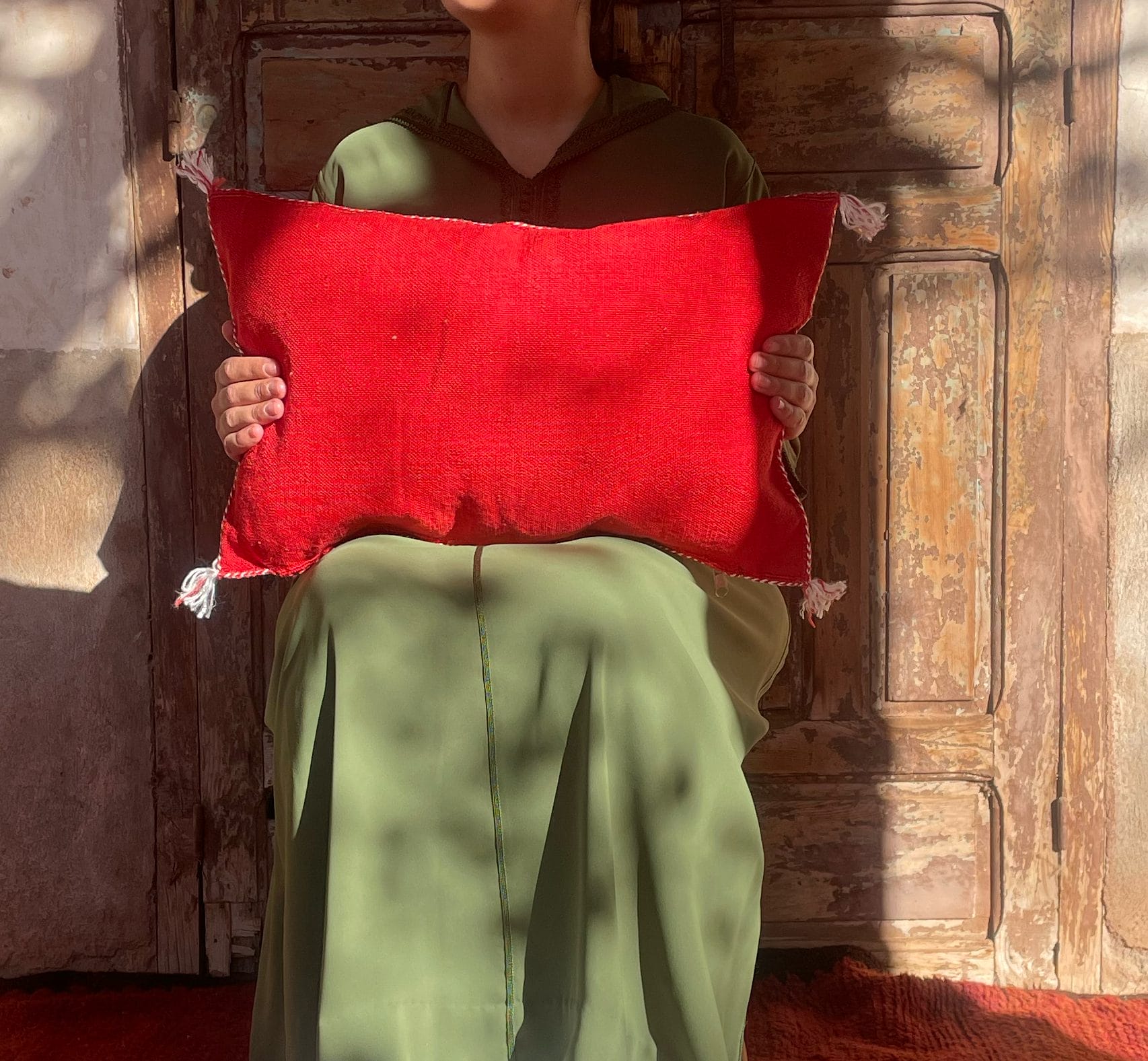 Een persoon in een groene outfit houdt een rood kussen vast terwijl hij voor een houten deur zit. Schaduwen en zonlicht vormen het decor.