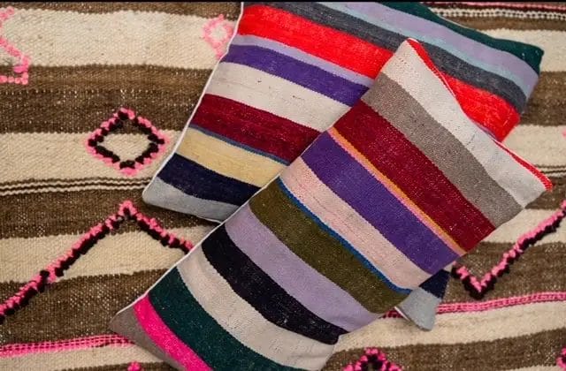 Twee levendige Kilim Vintage kussens - kleurrijk zijn geplaatst op een patroon vloerkleed met ruit- en lijnontwerpen. De kussens tonen horizontale strepen in een spectrum van kleuren zoals rood, paars, roze en groen.
