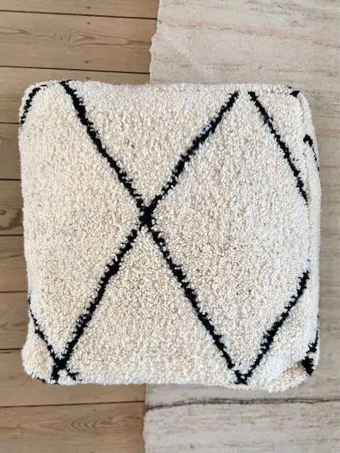 Een zachte, vierkante, gebroken witte poef met zwarte ruitpatronen, geplaatst op een houten vloer naast een beige tapijt.