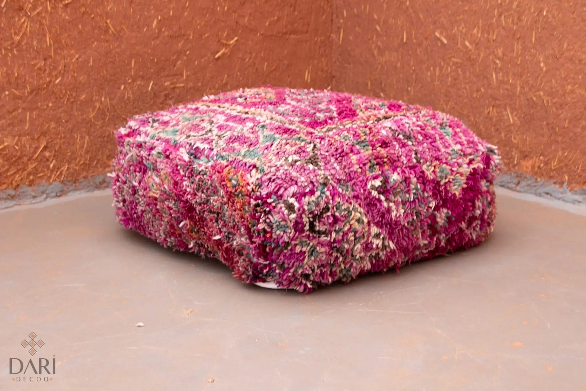 Een kleurrijke, gestructureerde poef met roze, rode en paarse patronen staat op een gladde, grijze vloer tegen een warme, bruine muur.