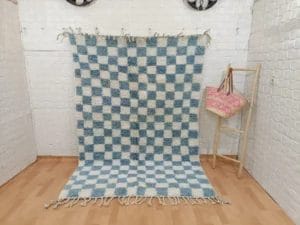 Een prachtig vervaardigd "100% HANDGEMAAKTE MAROKKAANSE TAPIJTEN TAPIJT 150 - 250 cm nr 189" met blauw-wit geblokte patronen en franjeranden hangt tegen een onberispelijk witte betegelde muur; een roze patroondoek drapeert elegant over een houten standaard aan de rechterkant.