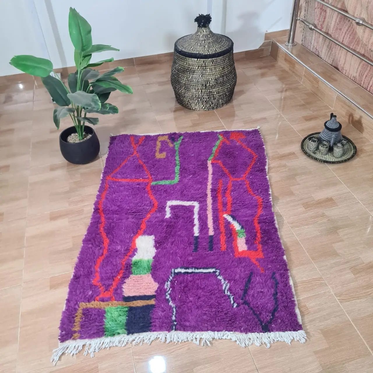Een 100% handgemaakte Marokkaanse tapijt, 150x100 cm (nr 133), met abstracte rode, groene, witte en zwarte patronen ligt op een gepolijste tegelvloer. Een potplant, geweven mand en decoratief item zijn eromheen geplaatst.