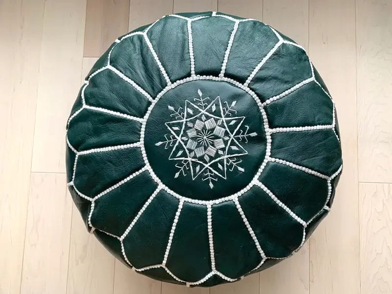 Een ronde, donkergroene poef met witte stiksels en een ingewikkeld geometrisch patroon op de bovenkant, geplaatst op een lichte houten vloer.
