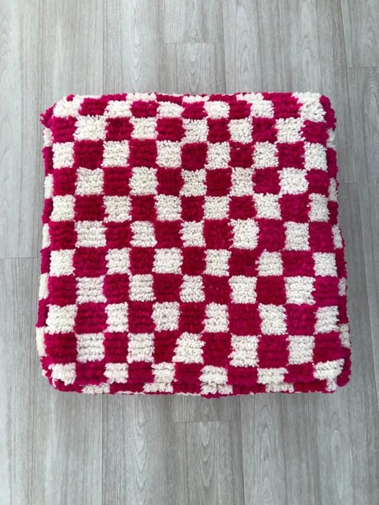 Een vierkant, rood-wit geblokt kussen ligt op een houten vloer naast een gezellige Marokkaanse Berber poef, roze.