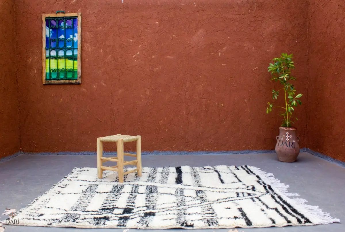 Een kamer met bruine muren met structuur, een kleurrijk raam, een klein krukje op een zwart-wit tapijt en een potplant in een pot met het Dari-logo.
