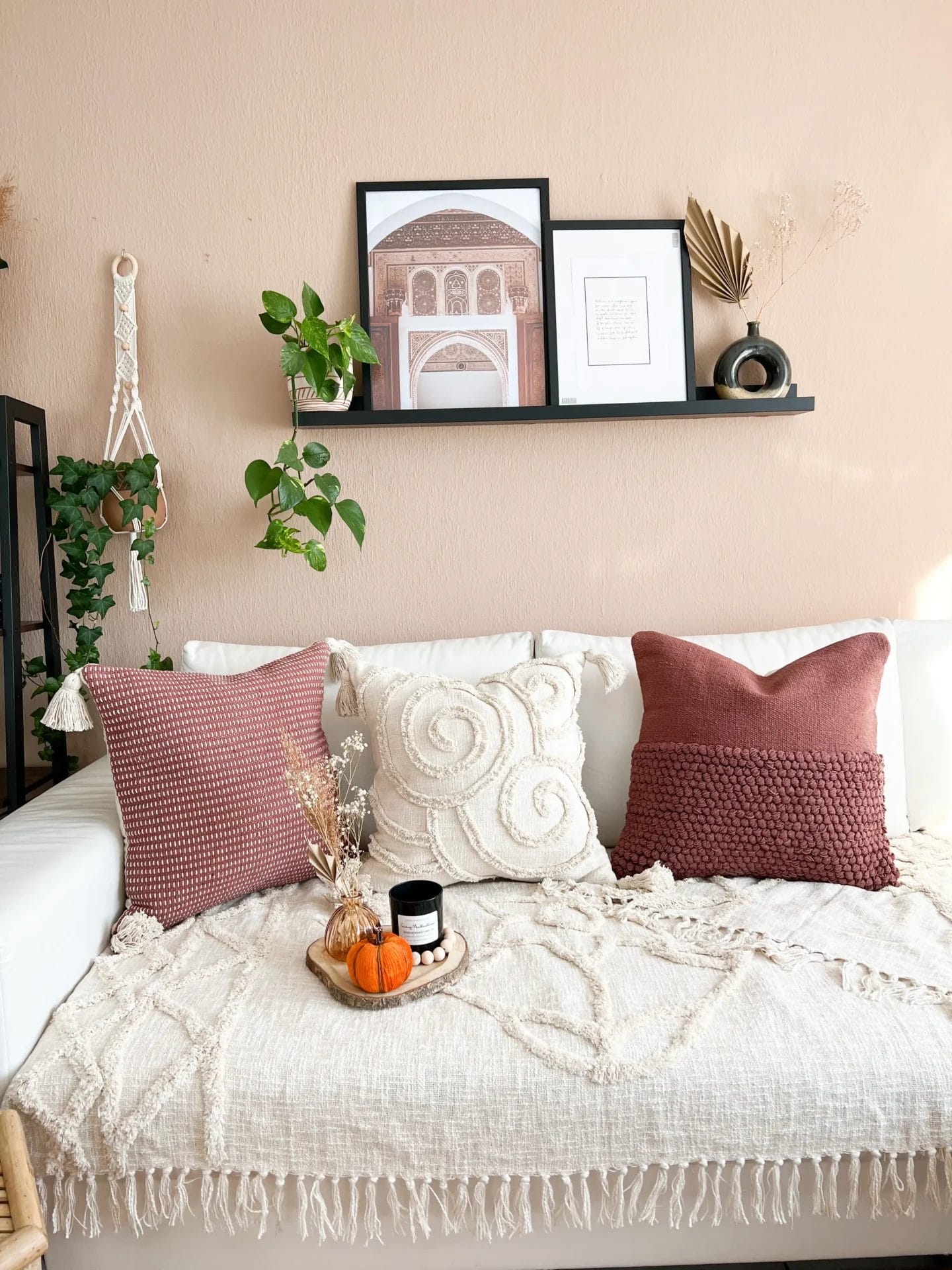Een gezellige woonkamer met een witte bank met kussens met textuur en een deken. Erboven is een plank met ingelijste kunst en planten. Een dienblad met een kaars en een kleine pompoen.