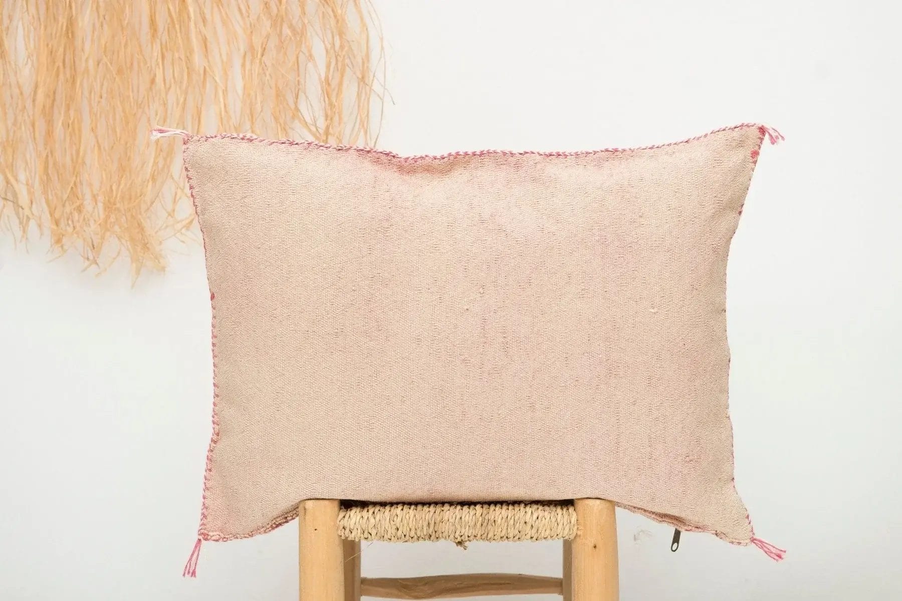 Een beige rechthoekig kussen met roze stiksels ligt op een geweven stoel tegen een witte muur. Een decoratief stro-element is gedeeltelijk zichtbaar aan de linkerkant.