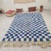 Een ruime 111 HANDGEMAAKT MAROKKAANSE_ BENI OUARAIN TAPIJTTAPIJT, met blauw-witte geblokte patronen en versierd met franjeranden, is elegant verspreid over een lichtgekleurde houten vloer. Kussens en een vaas zijn kunstig tegen de muur op de achtergrond geplaatst.