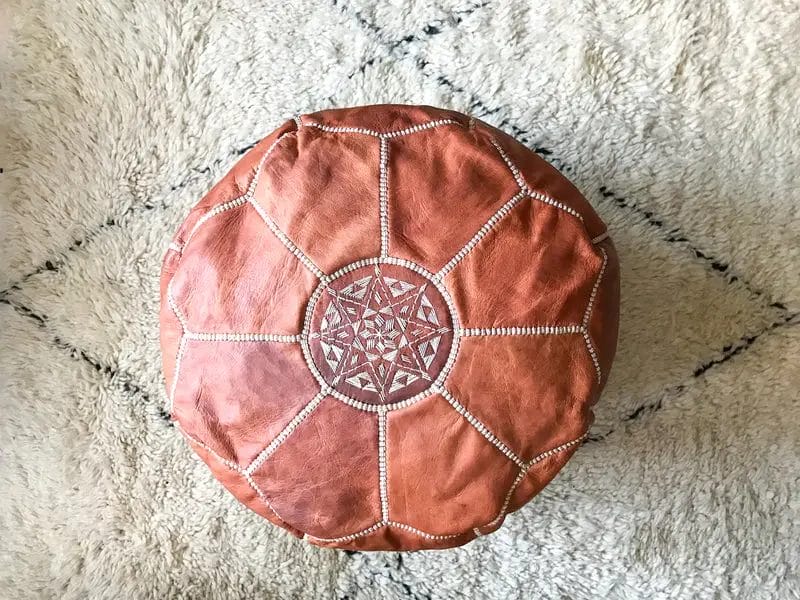 Een ronde, bruine leren poef met een geometrisch patroon op de bovenkant rust op een crèmekleurig, gestructureerd vloerkleed.