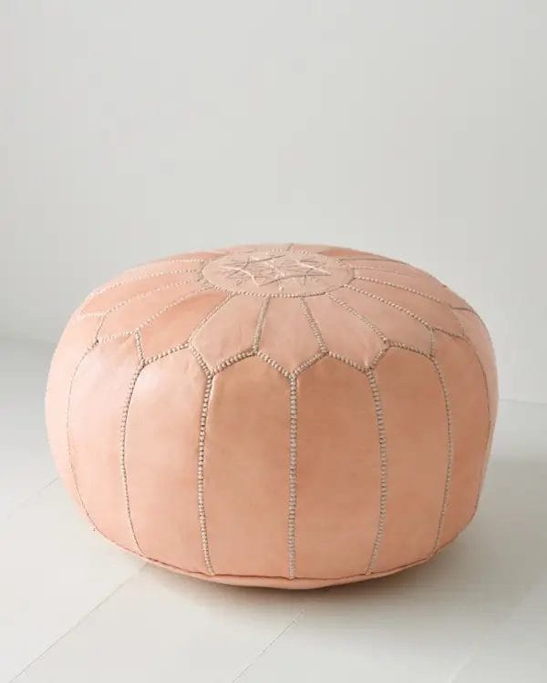 Een ronde, roze Marokkaanse leren voetenbank met gestikte decoratieve patronen staat op een witte vloer tegen een effen witte muur, aangevuld met een stijlvol vloerkleed.
