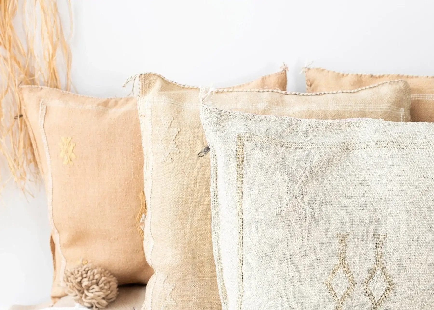 Vier beige en crèmekleurige sierkussens met geborduurde patronen zijn tegen een witte muur gestapeld, waardoor een gezellige sfeer ontstaat die perfect past bij de Houseify-esthetiek.